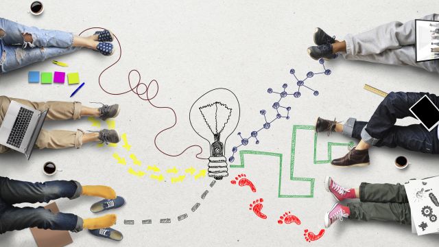Arbeiten bei der GRS Sechs Menschen sitzen auf dem Boden. Von Ihren Füßen sind unterschiedliche, bunte Fußspuren auf den Boden gemalt, die zu einer gemalten Glühbirne führen.