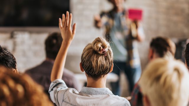 Rückenbild einer Frau, die in einem Seminar eine Frage hat und die Hand hebt